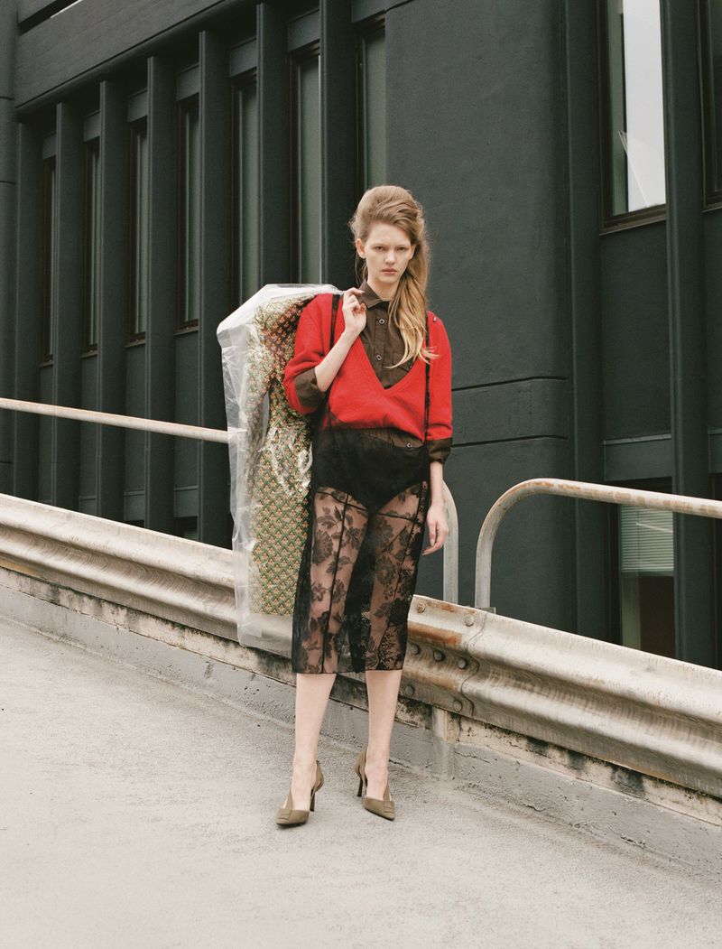 A blonde fashion model in a red sweater poses outdoors, holding a garment bag on her shoulder against an urban building backdrop.