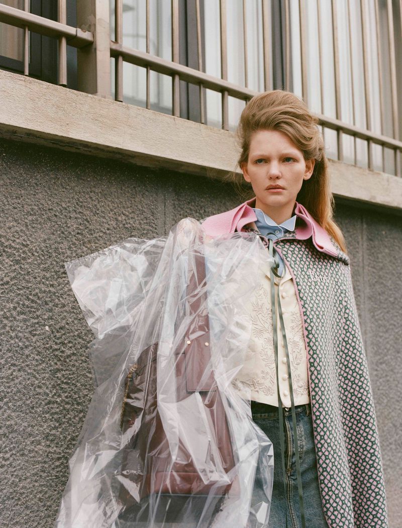 Fashionable adult female stands outdoors by a textured wall, carrying a garment bag with a brown handbag inside, wearing a patterned coat.