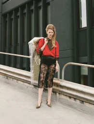 Model standing outdoors wearing a red sweater and black lace skirt, carrying a garment bag on a ramp