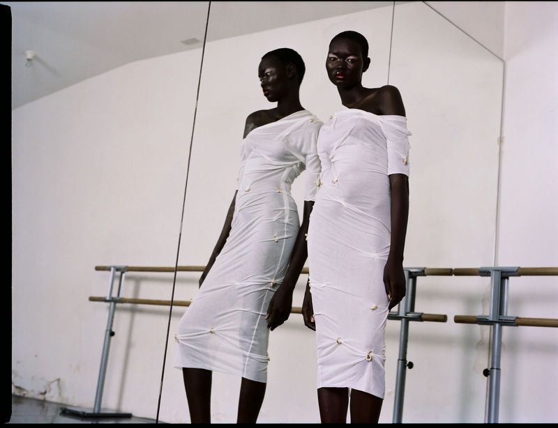 A Black female fashion model in a white dress poses in front of a mirror in a bright studio. The dress has decorative stitching.