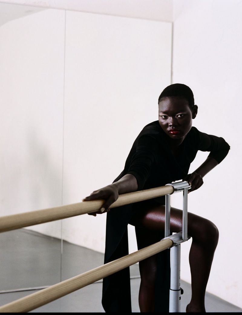 African woman poses at barre wearing black dress in dance studio. Mirror in background reflects bright light and a white wall.
