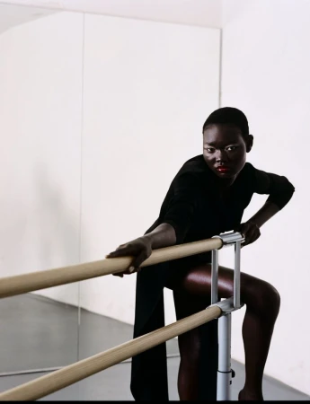 African woman poses at barre wearing black dress in dance studio. Mirror in background reflects bright light and a white wall.