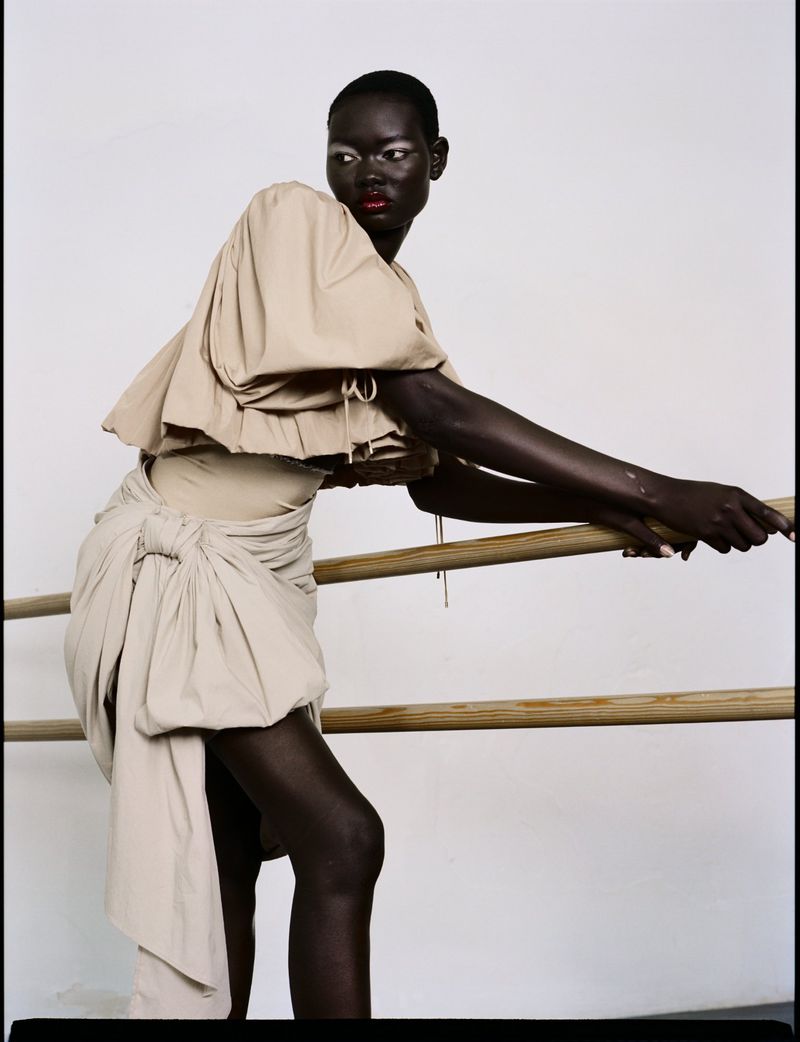 Fashion portrait of an African female model in a beige draped dress leaning against a barre in a white studio environment.