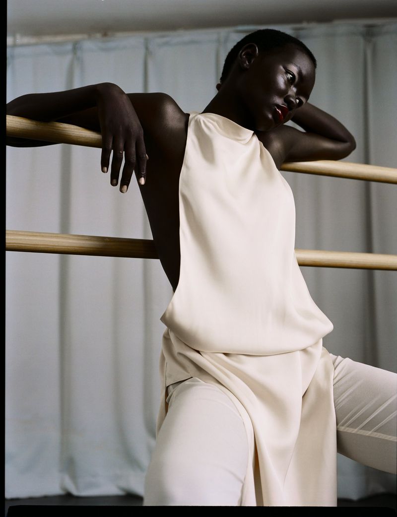 Dark-skinned woman in cream-colored clothing leaning on wood bar against a light blue curtain in portrait orientation, looking down.
