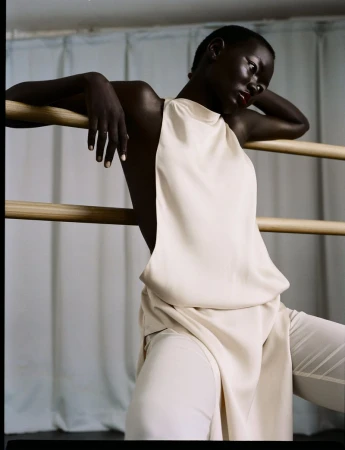 Dark-skinned woman in cream-colored clothing leaning on wood bar against a light blue curtain in portrait orientation, looking down.