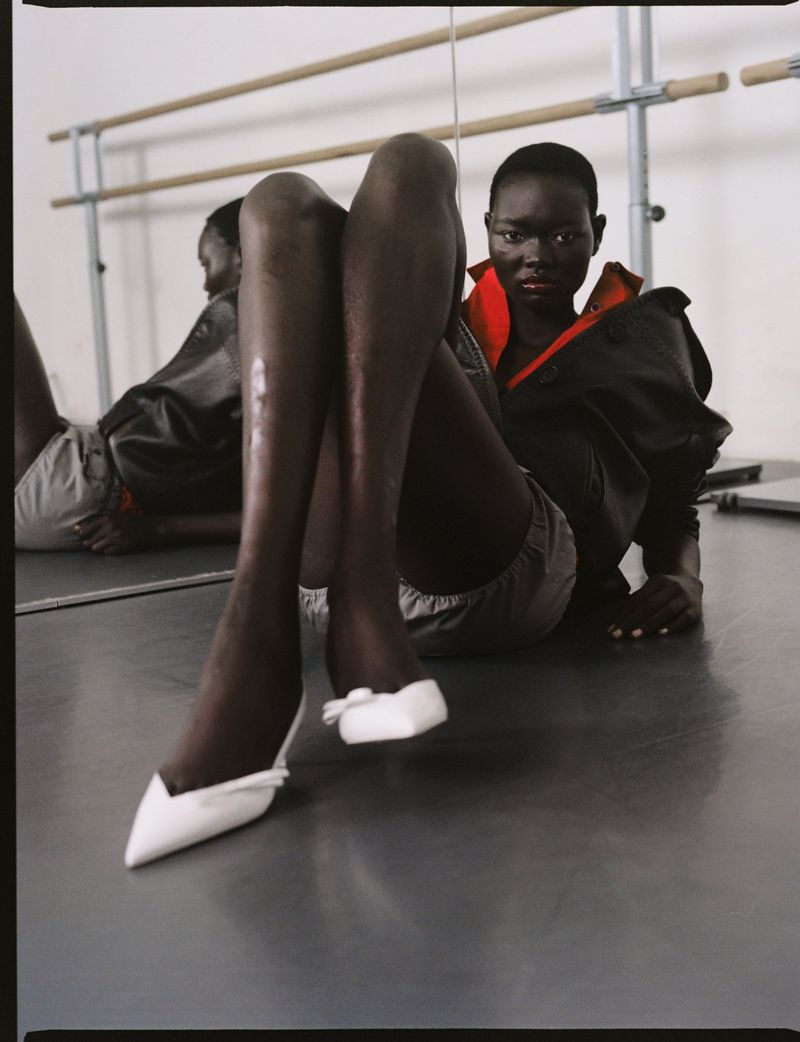 Black model wearing black jacket and white shoes seated in front of ballet barres in a dance studio