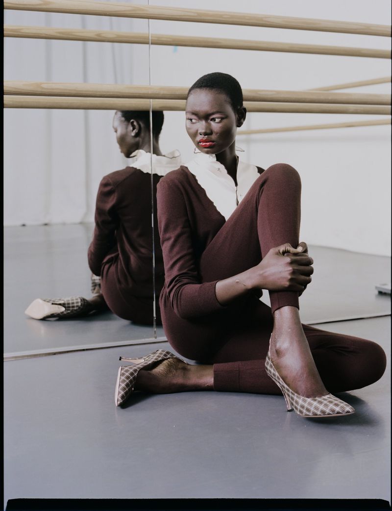 Elegant black woman in brown knitwear and patterned heels poses in front of a mirror in a light-filled studio setting.