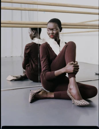 Elegant black woman in brown knitwear and patterned heels poses in front of a mirror in a light-filled studio setting.
