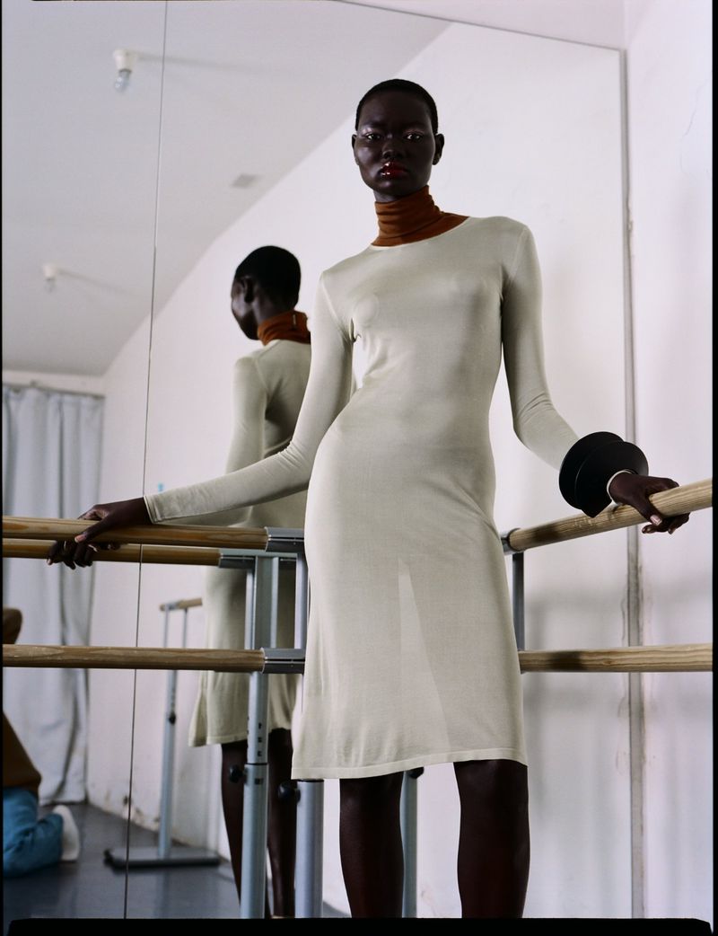 Elegant dark-skinned woman poses in long-sleeved beige dress in a dance studio with barre and mirror, showcasing fashion and style.