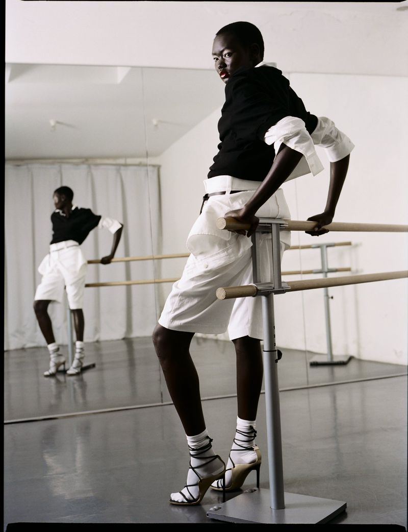 Fashion shot of an adult female with dark skin, wearing high heels, white shorts, and a black top, leaning on a ballet barre in a dance studio.