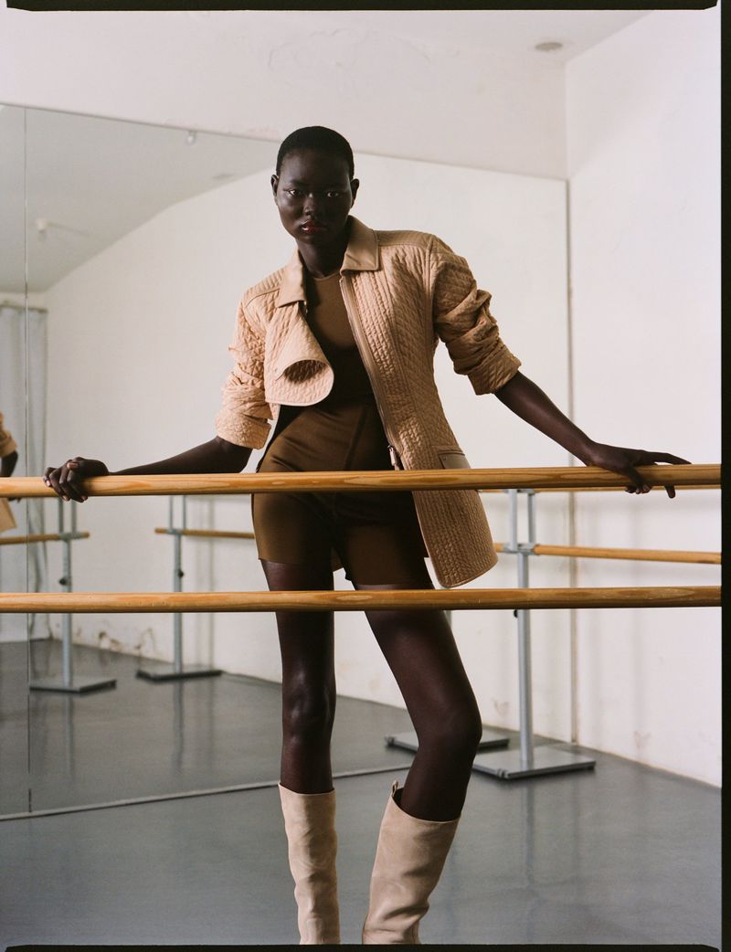 Adult woman in a beige jacket and matching knee-high boots posing at a barre in dance studio with a mirror and neutral lighting.