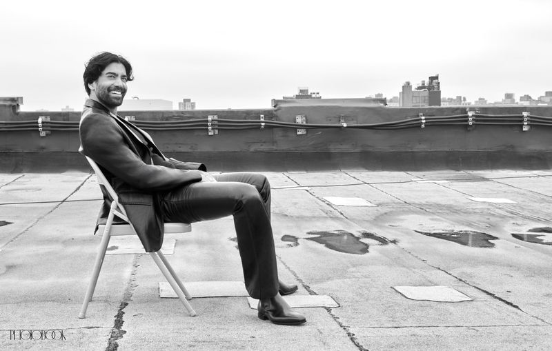 Monochrome shot of an adult man with dark hair, wearing a suit, sitting on a white chair on a rooftop with city buildings in background.
