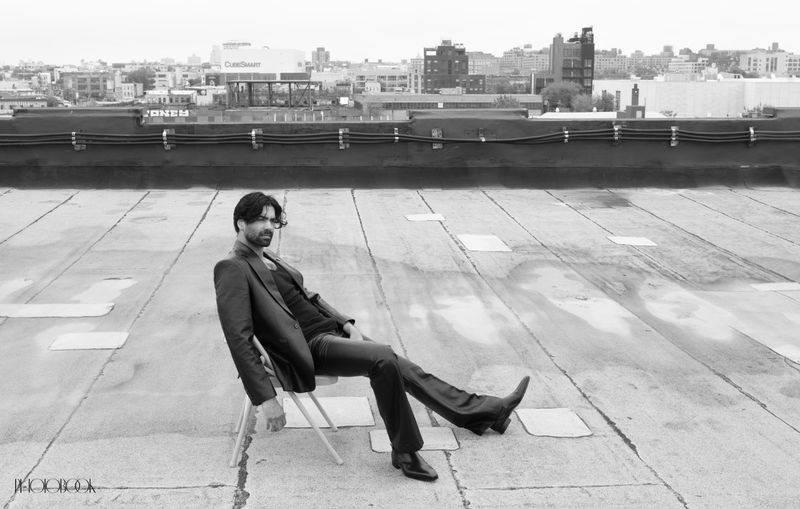 Black and white portrait of an adult man in a suit sitting on a chair on a rooftop overlooking a cityscape with a cloudy sky.