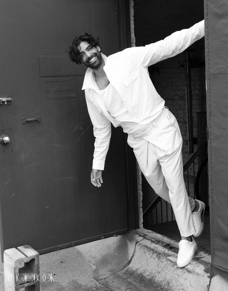Black and white studio portrait of a smiling adult male fashion model posing in white casualwear and sneakers at a doorway.