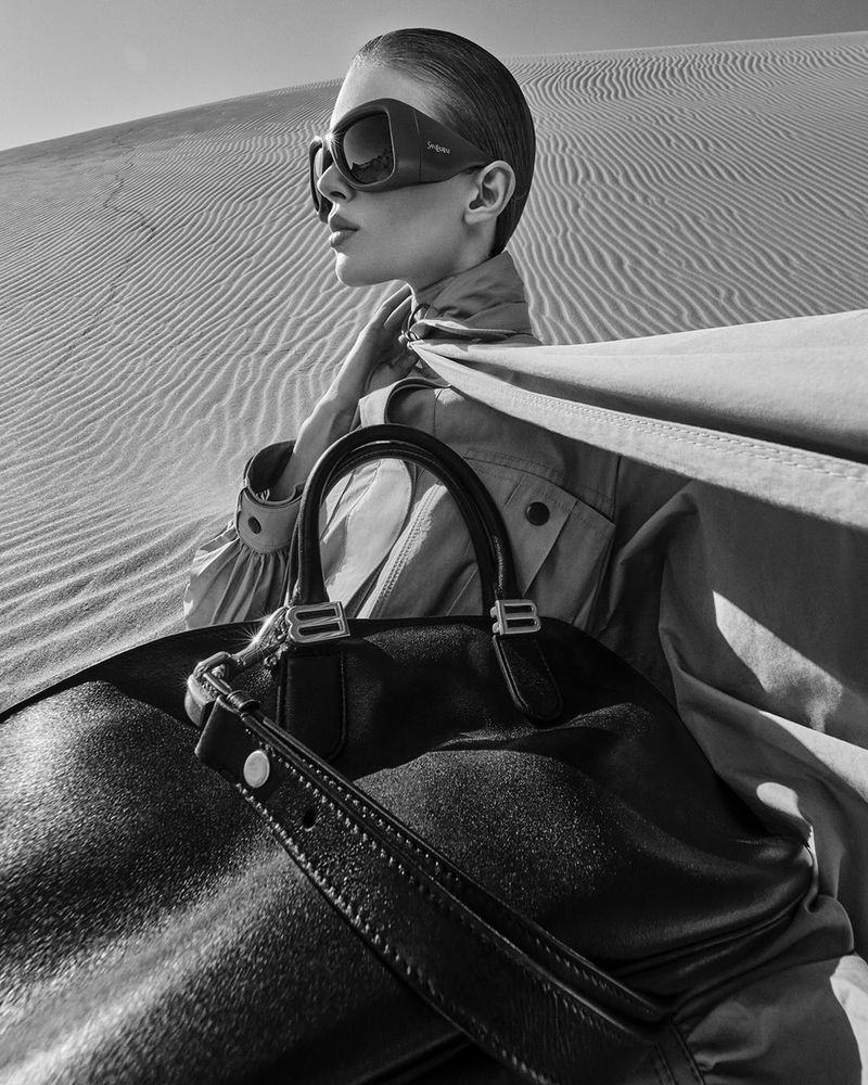 Monochrome fashion shot of a woman in sunglasses and a trench coat with a purse, posing in a desert landscape with sand dunes.
