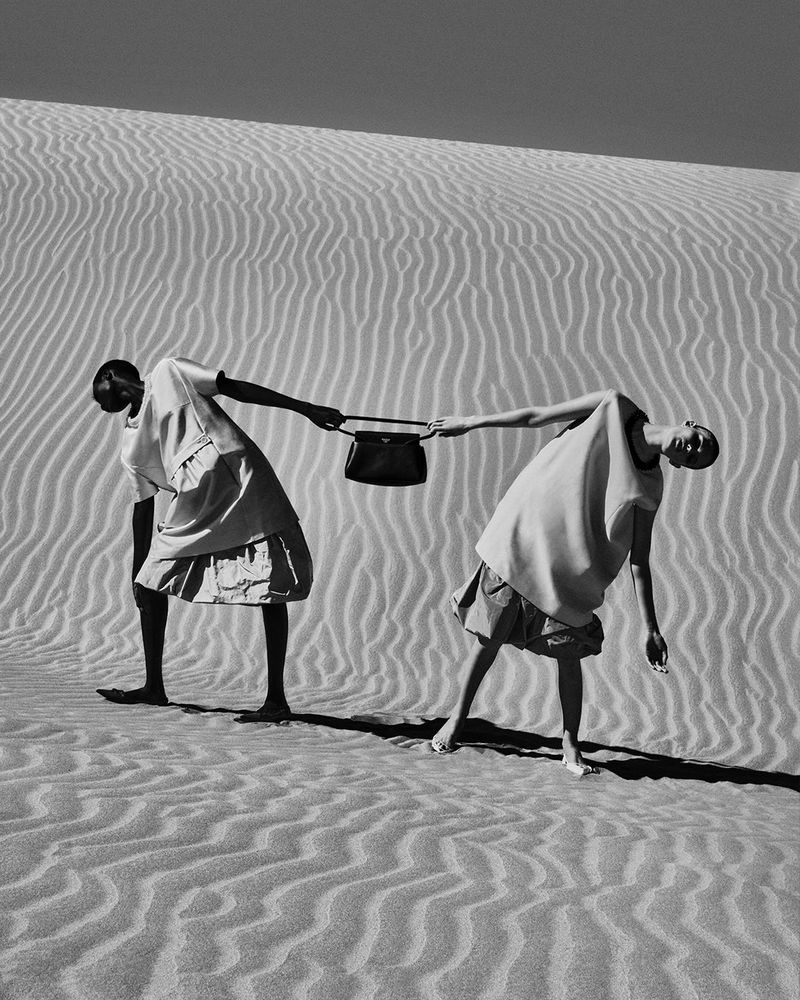 Black and white fashion shot of two women on a sand dune holding a handbag, featuring minimalistic dresses and contrasting dark skin tones.