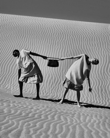 Black and white fashion shot of two women on a sand dune holding a handbag, featuring minimalistic dresses and contrasting dark skin tones.