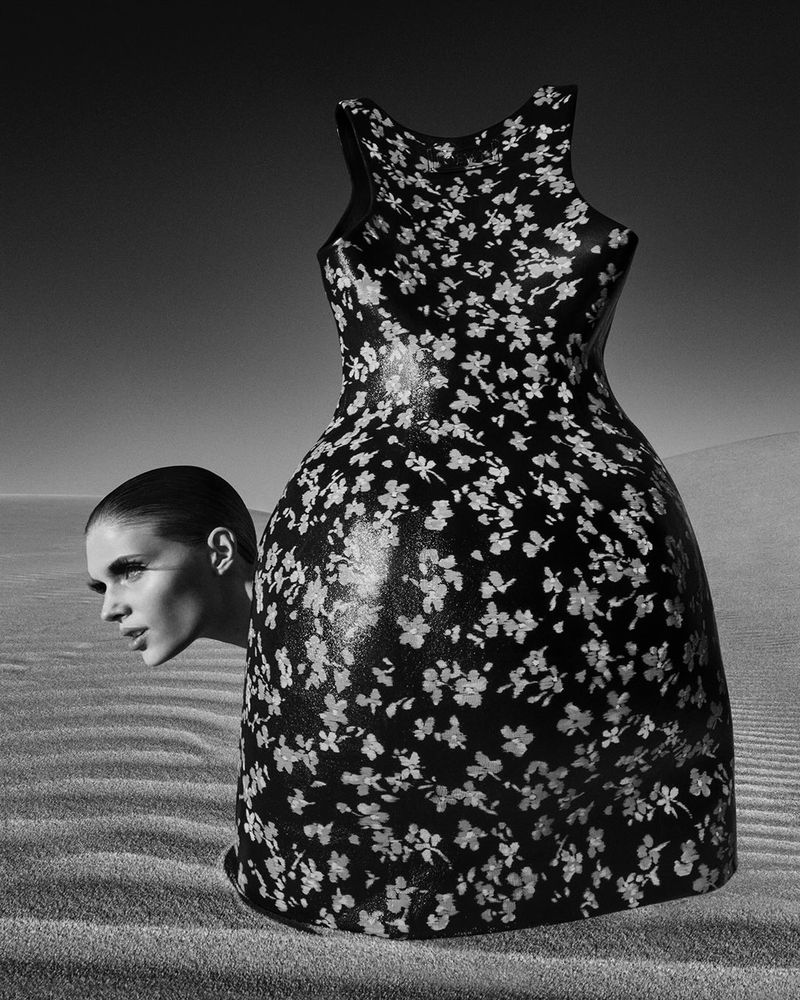 Black and white fashion image featuring a woman's head emerging from a floral dress against a sand dune background under a clear sky.