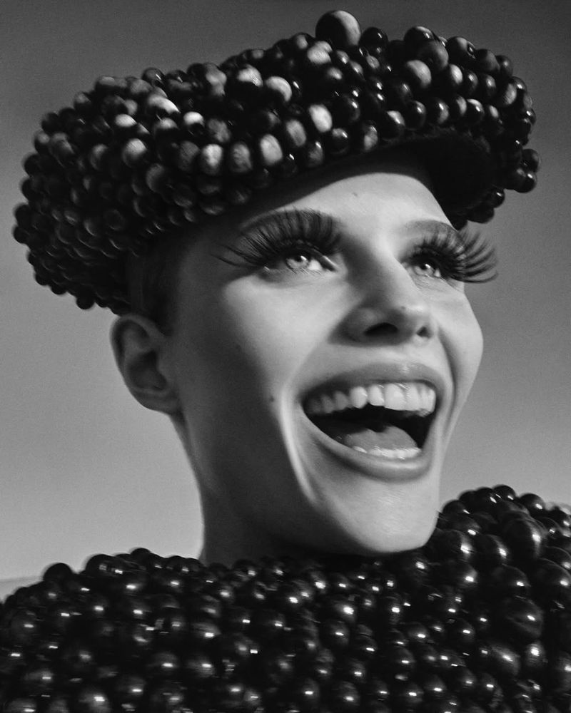 Monochrome portrait of smiling female with exaggerated eyelashes and headpiece, chestpiece covered in black spheres against gray backdrop.