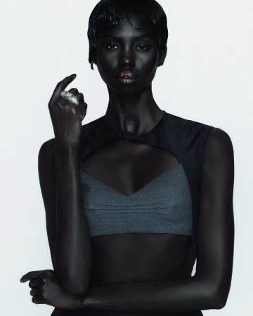 Close-up portrait of a Black female adult model in a gray top with a cut-out design, against a white backdrop, with a neutral facial expression.