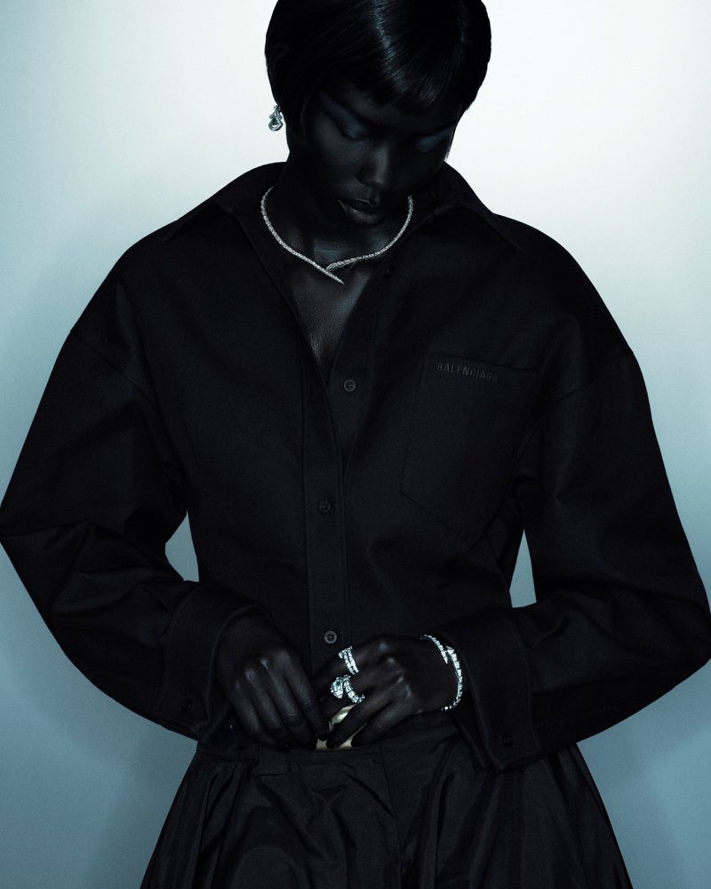 Woman in a black oversized shirt adjusting a skirt with jewelry details including a necklace, earrings, rings, and a bracelet.