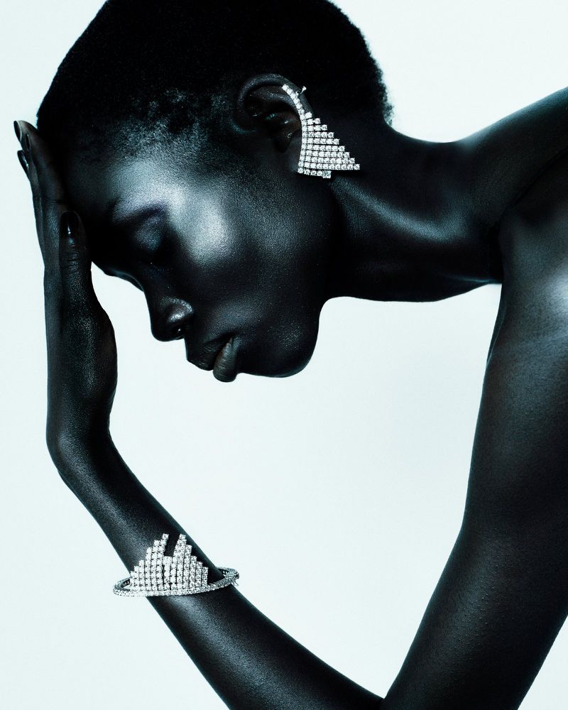 Elegant close-up of a dark-skinned woman with a striking triangular earring and jeweled bracelet, set against a clean, light background.