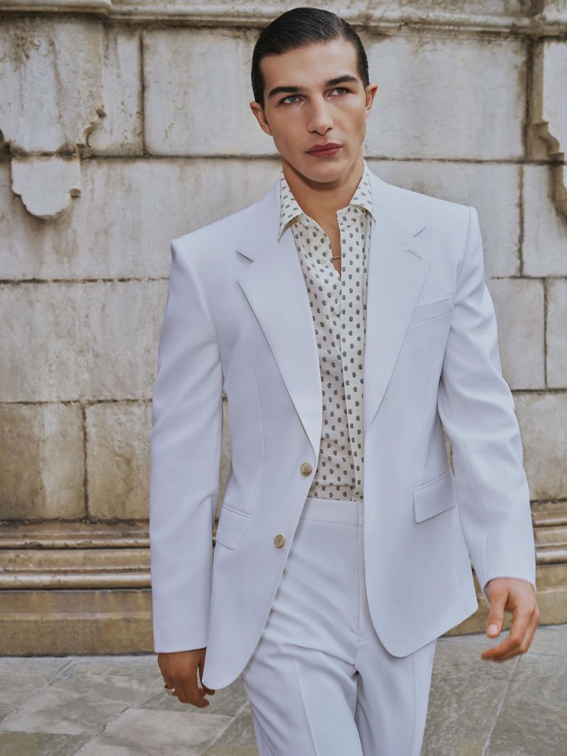 Man wearing a light suit jacket and patterned shirt standing outdoors in front of a stone wall.