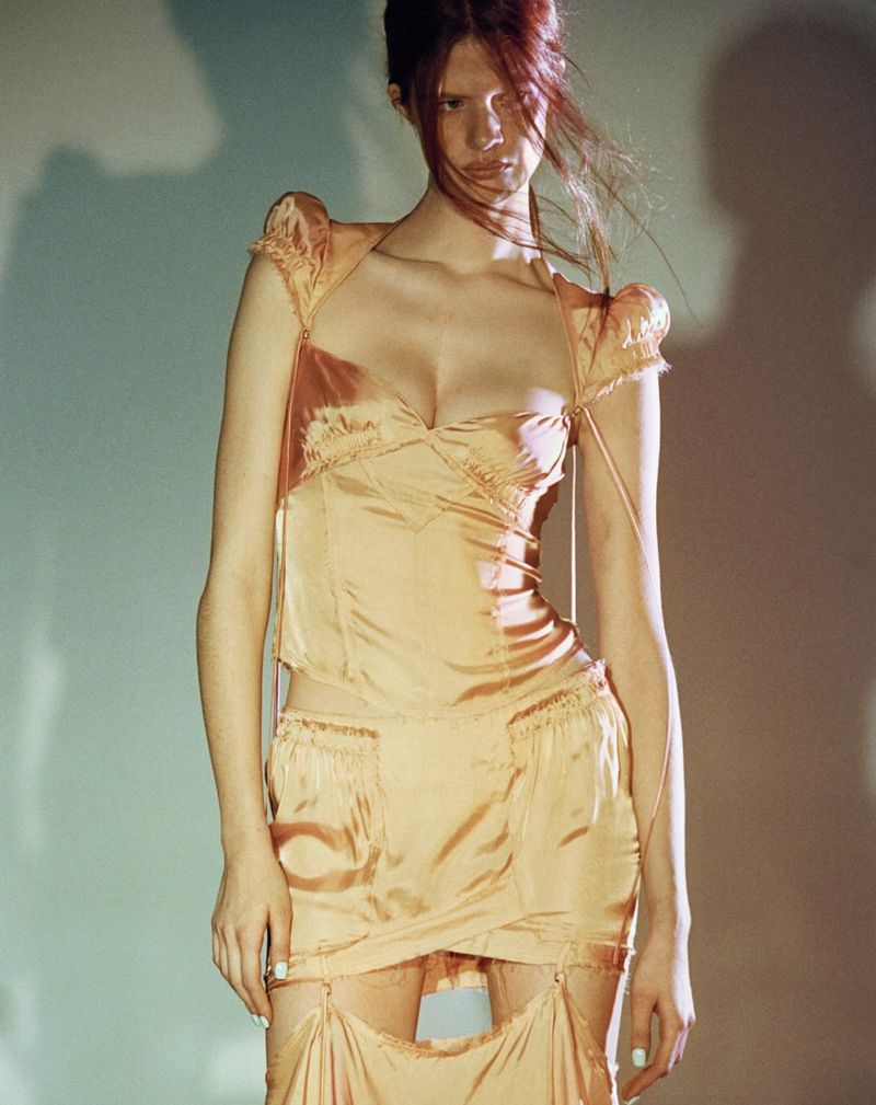 A female fashion model poses in a draped peach dress in a studio with soft lighting, showcasing modern editorial style and design.