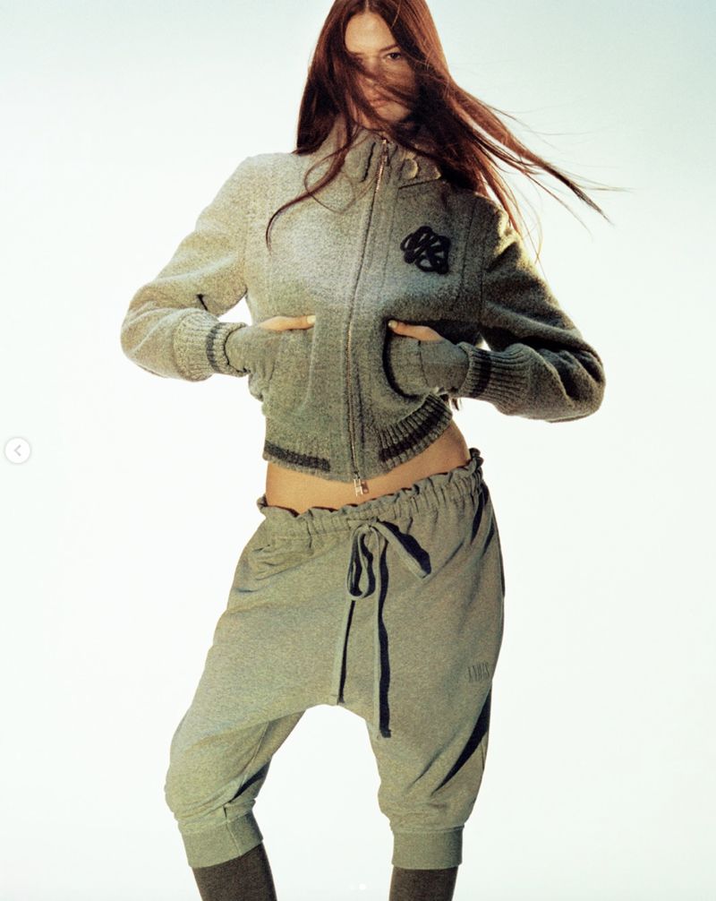 Adult female fashion model poses in a grey sweater and sweatpants against a gradient white backdrop in a studio setting.