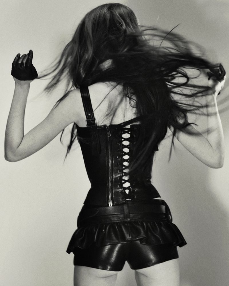 Back view of a slender woman with long hair, wearing a black leather corset, shorts, and gloves, arms raised against a grey background.