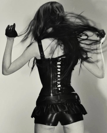 Back view of a slender woman with long hair, wearing a black leather corset, shorts, and gloves, arms raised against a grey background.