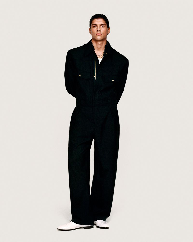 Full-length studio shot of a young adult male in a black jumpsuit and white shoes, with hands behind his back against a white background.