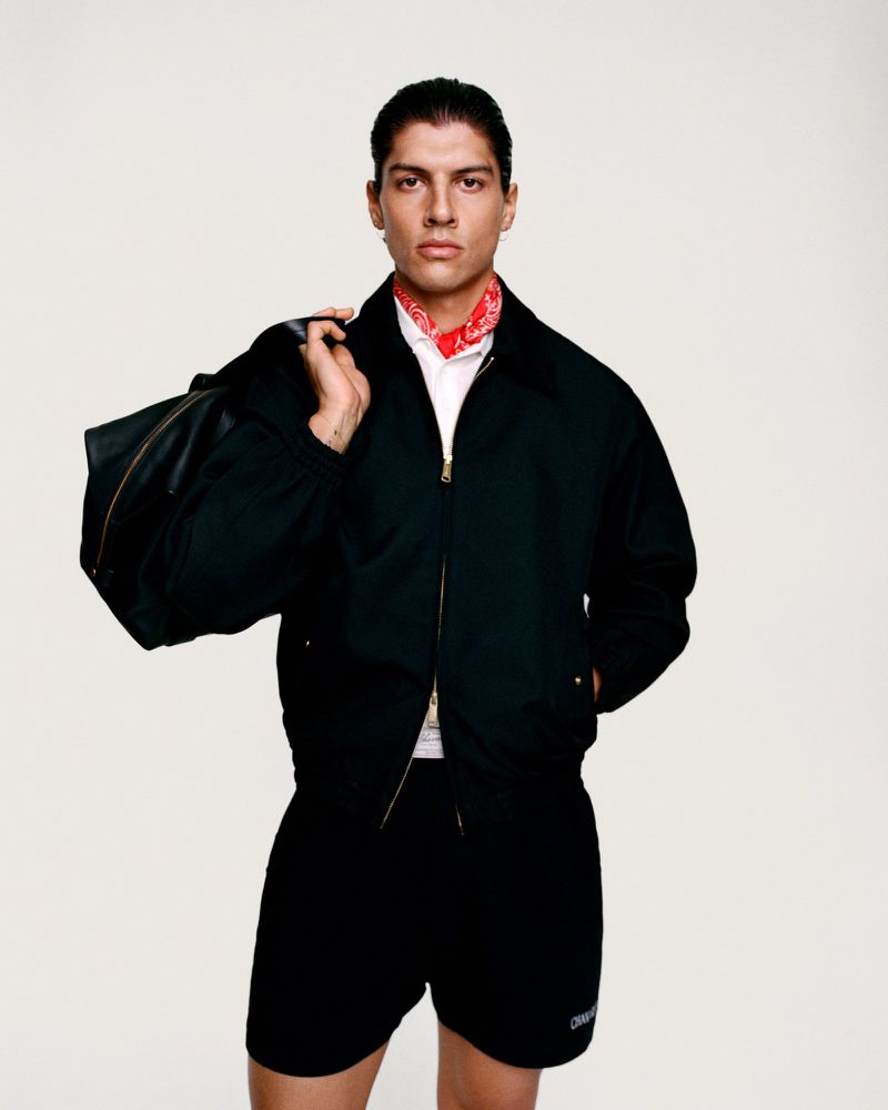 Adult male in a black jacket and shorts carries a duffel bag in a studio shot, accessorized with a red bandana around the neck.