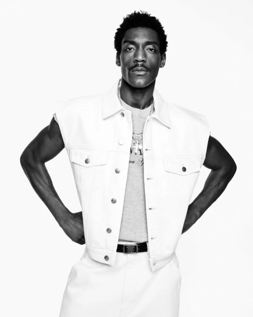 Black and white portrait of an adult male in a white denim vest and pants with a grey tee shirt posing in a photo studio.