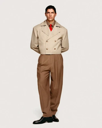 Full-length shot of a young adult male in a beige cropped double-breasted jacket, brown dress pants, shiny black shoes, and a red tie on a white background.