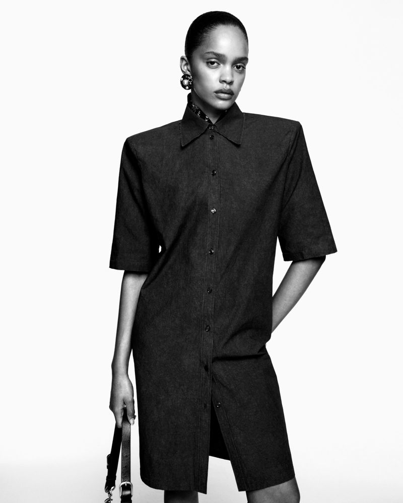 Black and white studio shot of a young adult woman in a collared, button-down dress, holding a purse with statement earrings.