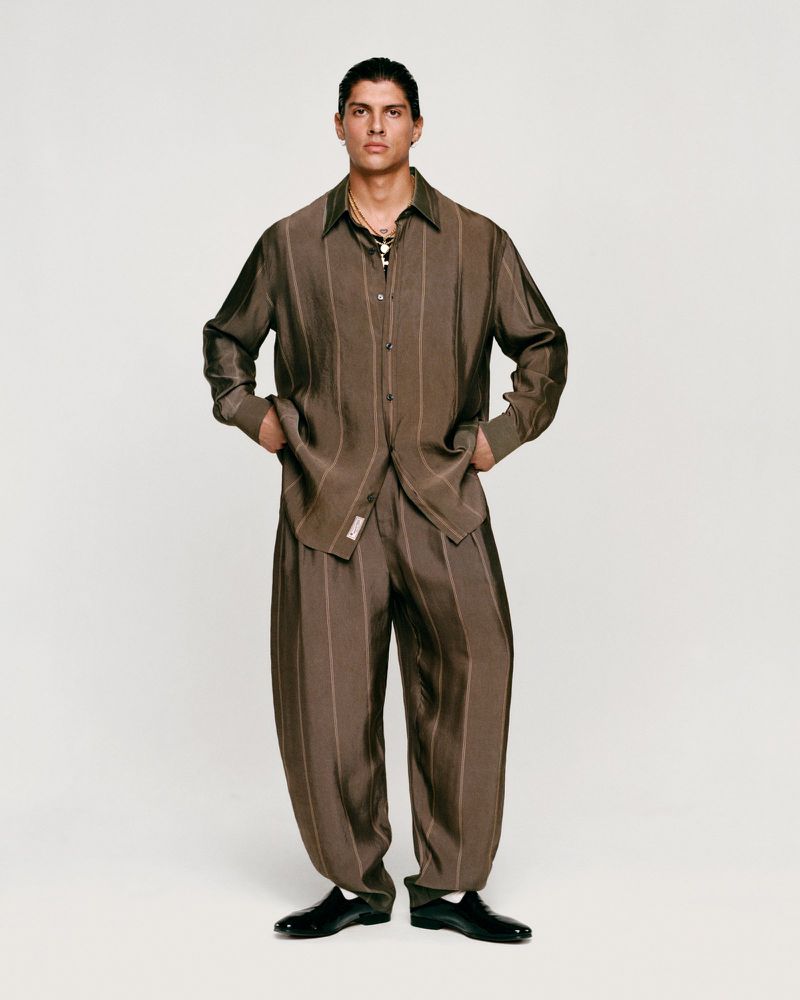 Male model poses in a brown striped shirt and baggy pants against a neutral background, showcasing a fashion editorial style with natural light.