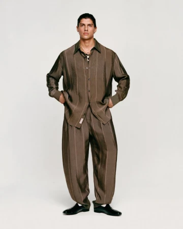 Male model poses in a brown striped shirt and baggy pants against a neutral background, showcasing a fashion editorial style with natural light.