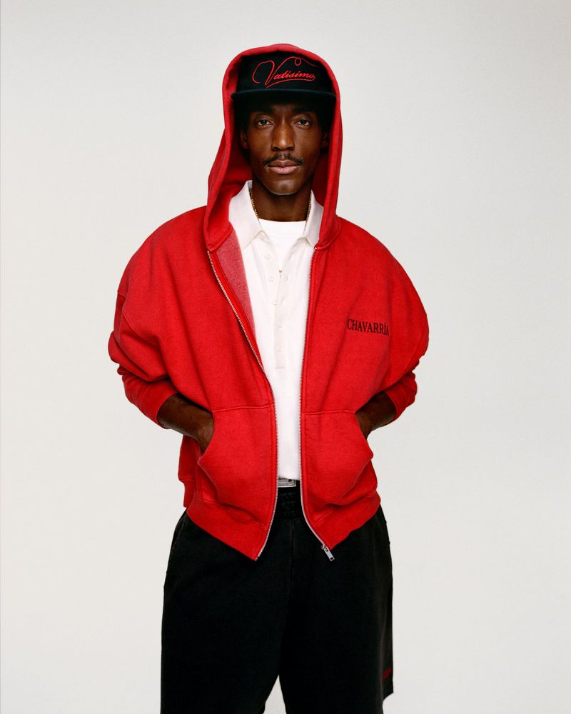 Portrait of a young African American male wearing a red hooded sweatshirt and a black baseball cap, posed against a white backdrop.
