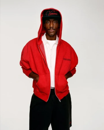 Portrait of a young African American male wearing a red hooded sweatshirt and a black baseball cap, posed against a white backdrop.