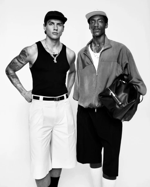 Two men pose in a black and white studio shot. One man wears a tank top and hat, the other wears a jacket and carries a bag.