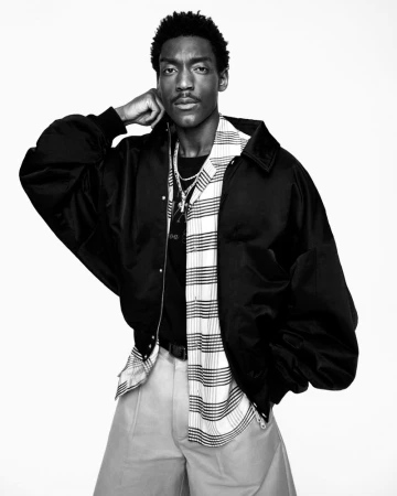 Black man wearing oversized black jacket, striped open shirt, and light trousers against white backdrop
