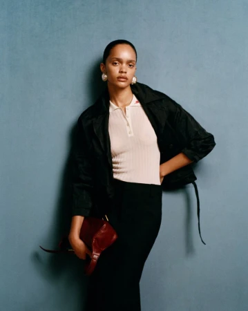 A woman stands against a blue wall, wearing a white ribbed polo shirt, a black jacket, and black trousers, holding a dark red leather bag.