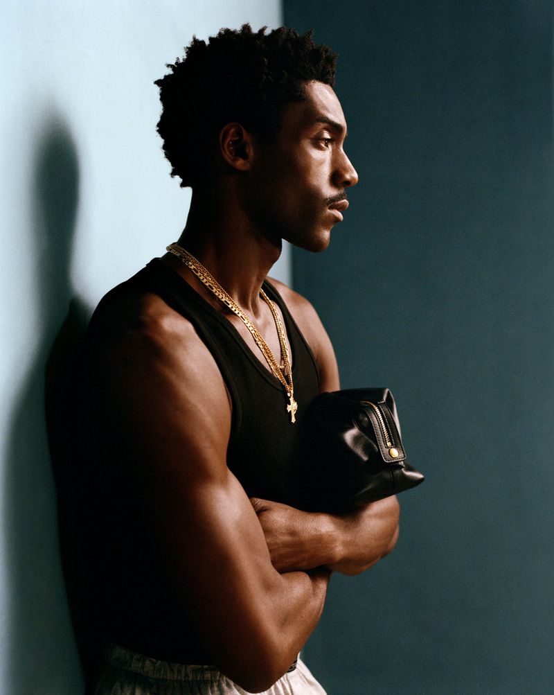 Profile portrait of a young adult man with a muscular build, wearing a black tank top and gold chains, holding a black bag indoors.
