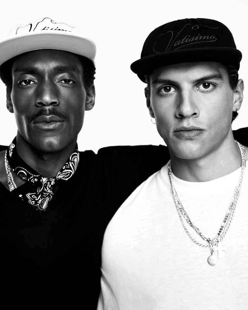 Monochrome portrait of two adult men wearing baseball caps and casual attire, embracing with a light background in a photo studio.