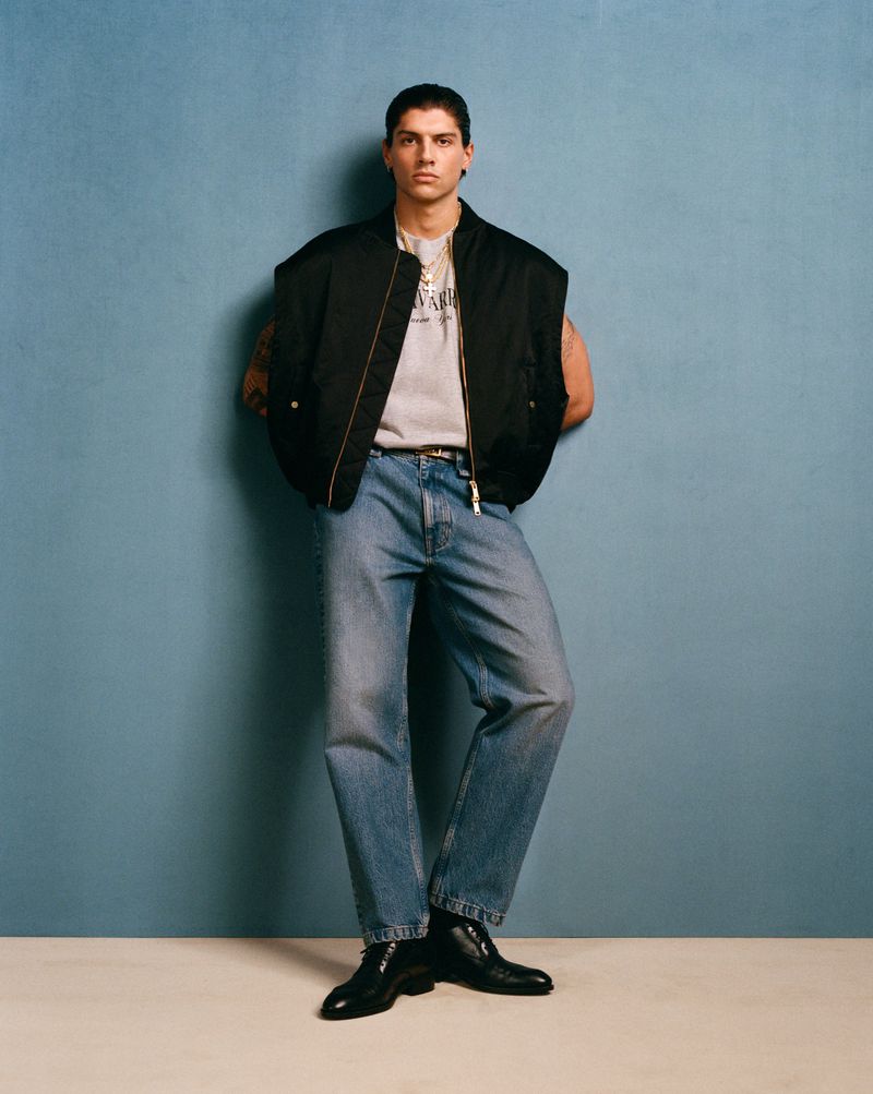 Full-length shot of a young adult man in a black vest, graphic tee, blue jeans, and black boots against a blue backdrop.