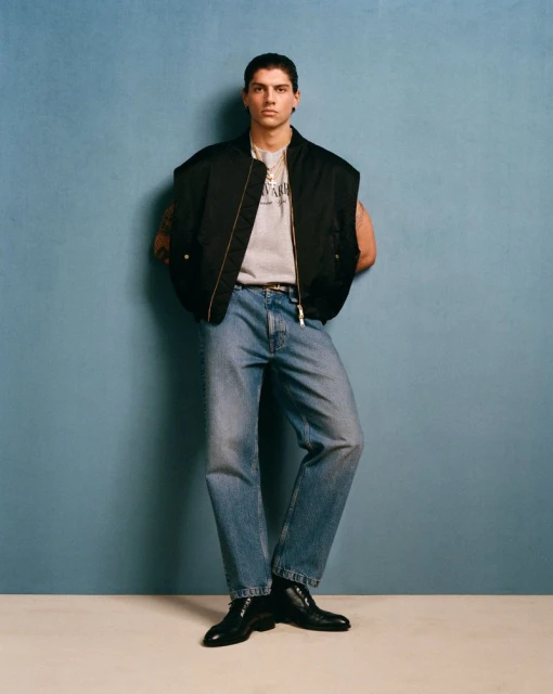 Full-length shot of a young adult man in a black vest, graphic tee, blue jeans, and black boots against a blue backdrop.