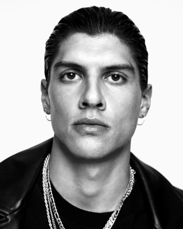 Monochrome portrait of a handsome adult male with slicked-back hair, gold hoop earrings, and silver chain necklaces against white backdrop.