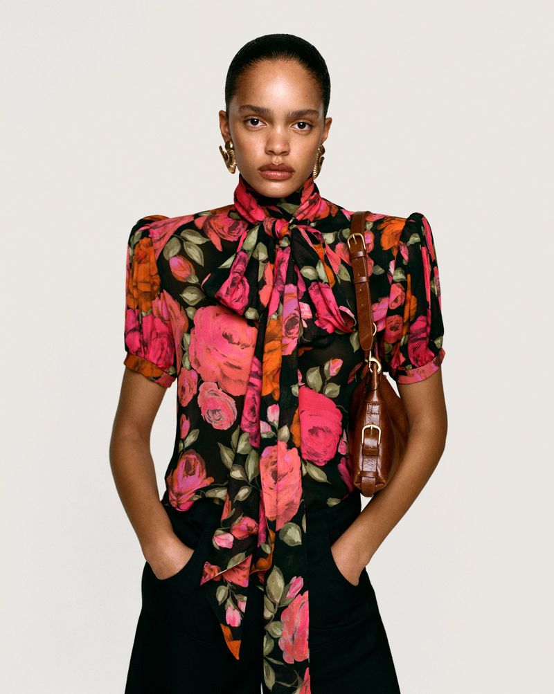 A woman models a floral blouse with a neck tie and a brown purse against a neutral backdrop, showcasing fashion and style.