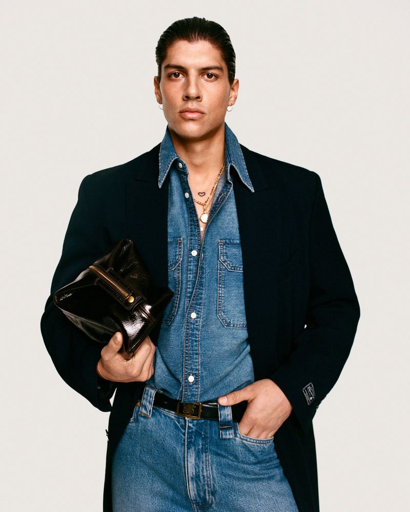 Fashionable adult male in denim shirt and jeans, holding a dark glossy clutch bag, posing against a white backdrop in a studio setting.